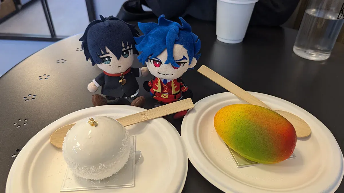 Tadashi and Ainosuke puppet charms sitting next to two small cakes. One of the small cakes is white and smooth, while the other is shaped like a ripe mango with the skin still on.