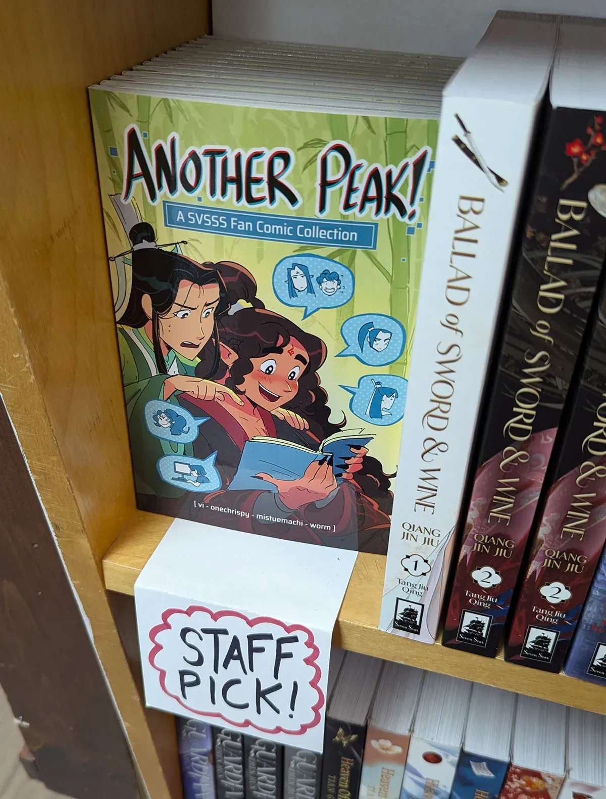 A copy of the fancomic "Another Peak: a SVSSS Fan Comic Collection" by the cartoonists vi, onechrispy, Mitsuemachi, worm sits next to a stack of danmei novels with the label "Staff Pick" written in all caps under the fancomic